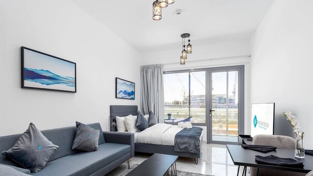 A modern hotel room with a grey sofa, a bed with grey bedding, a desk with a vase of flowers, and a large window with a city view. - Bloomfields New STU in Oasis Masdar City (Abu Dabi)
