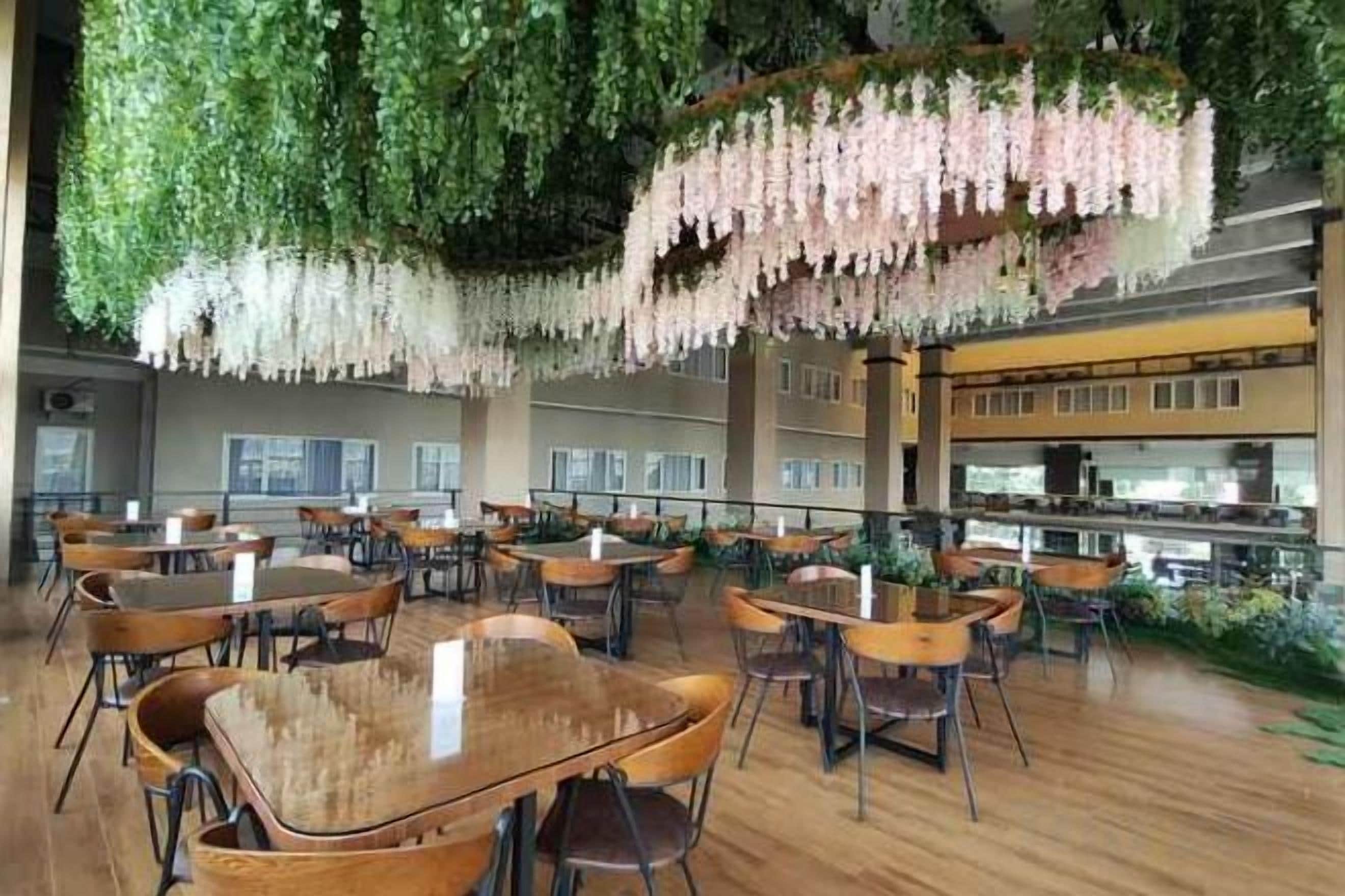 Een ruime, overdekte eetruimte met meerdere tafels en stoelen, een hoog plafond met hangende beplanting, en een zicht op de keuken in de achtergrond.