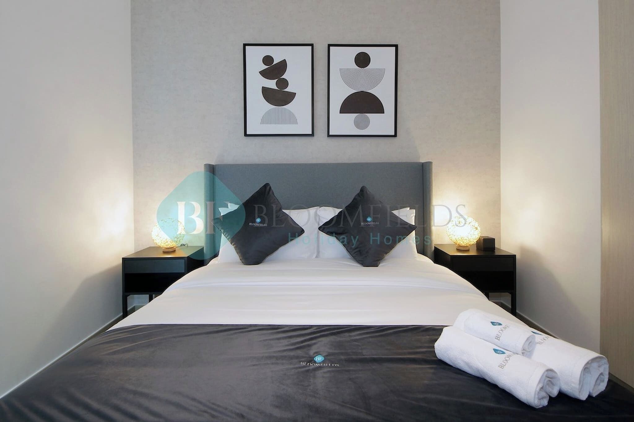A hotel room with a bed, two pillows, a bedside table with a lamp, and two framed artworks on the wall.