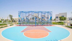 Una cancha de baloncesto al aire libre con superficie azul y naranja, rodeada por una valla azul con un aro de baloncesto.
