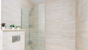Une douche fermée par des parois en verre, dotée d’un pommeau de douche à main et d’un robinet à main. Trois plantes en pot sont disposées sur une étagère à gauche.