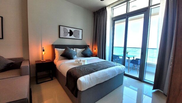 A modern hotel room with a large bed, a nightstand with a lamp, a sofa, and a view of the sea through large windows. - Bloomfields STU with Canal View Julphar Al Reem (Abu Dabi)