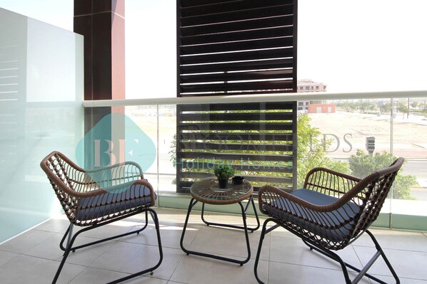 A balcony with wicker chairs and a small round table, a view of a building, and a fence in the background.