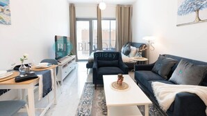Una sala moderna con sofá, mesa de centro y televisor. Cuenta con un balcón con cortinas y un cuadro de un árbol.