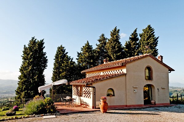 Un petit bâtiment d’un seul étage, doté d’un toit en tuiles, entouré de grands arbres et bordé d’un chemin de gravier.