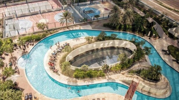 Et luftfoto viser en swimmingpool med et buet design, omgivet af palmer og liggestole.