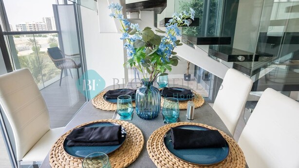 A dining table set with woven placemats, dark napkins, and blue glassware, with a vase of flowers in the center.