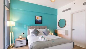 A modern bedroom with a teal accent wall, a bed with grey bedding, a bedside table with a lamp, and a framed picture above the bed.