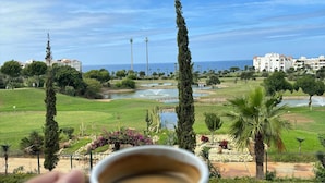 A hand holding a cup of coffee with a view of a landscaped garden, a pool, and buildings in the background. - For rent: Luxury apartment in a tourist residence. (Asilah)