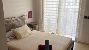 A neatly made bed with a patterned headboard, a red lamp, and a window with sheer curtains. - For rent: Luxury apartment in a tourist residence. (Asilah)