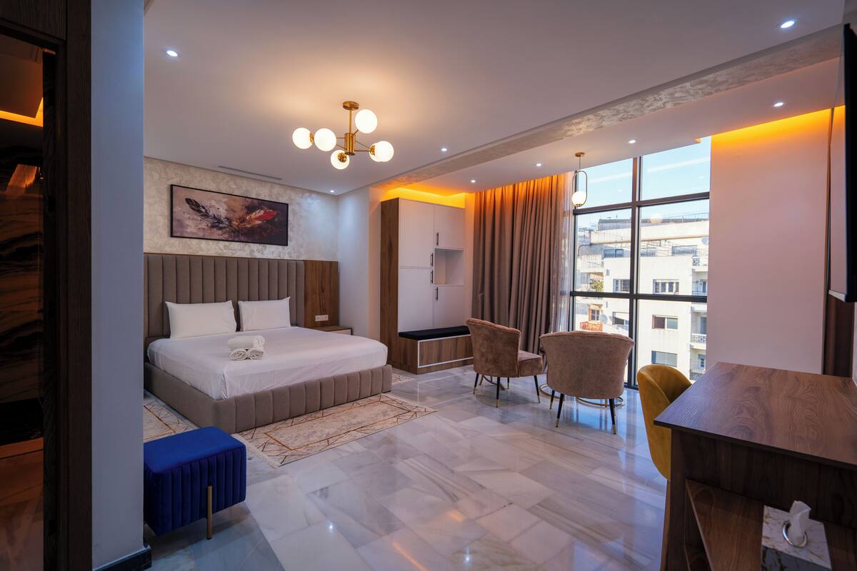 A modern hotel room with a large bed, a desk, and a chair. There is a window with curtains and a view of buildings outside.