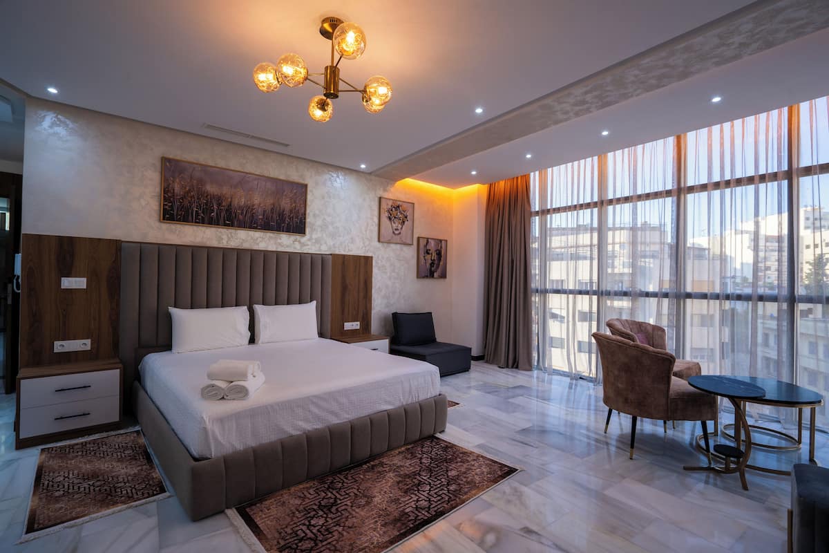 A modern hotel room with a large bed, a seating area with a table, and large windows offering a view of the city.