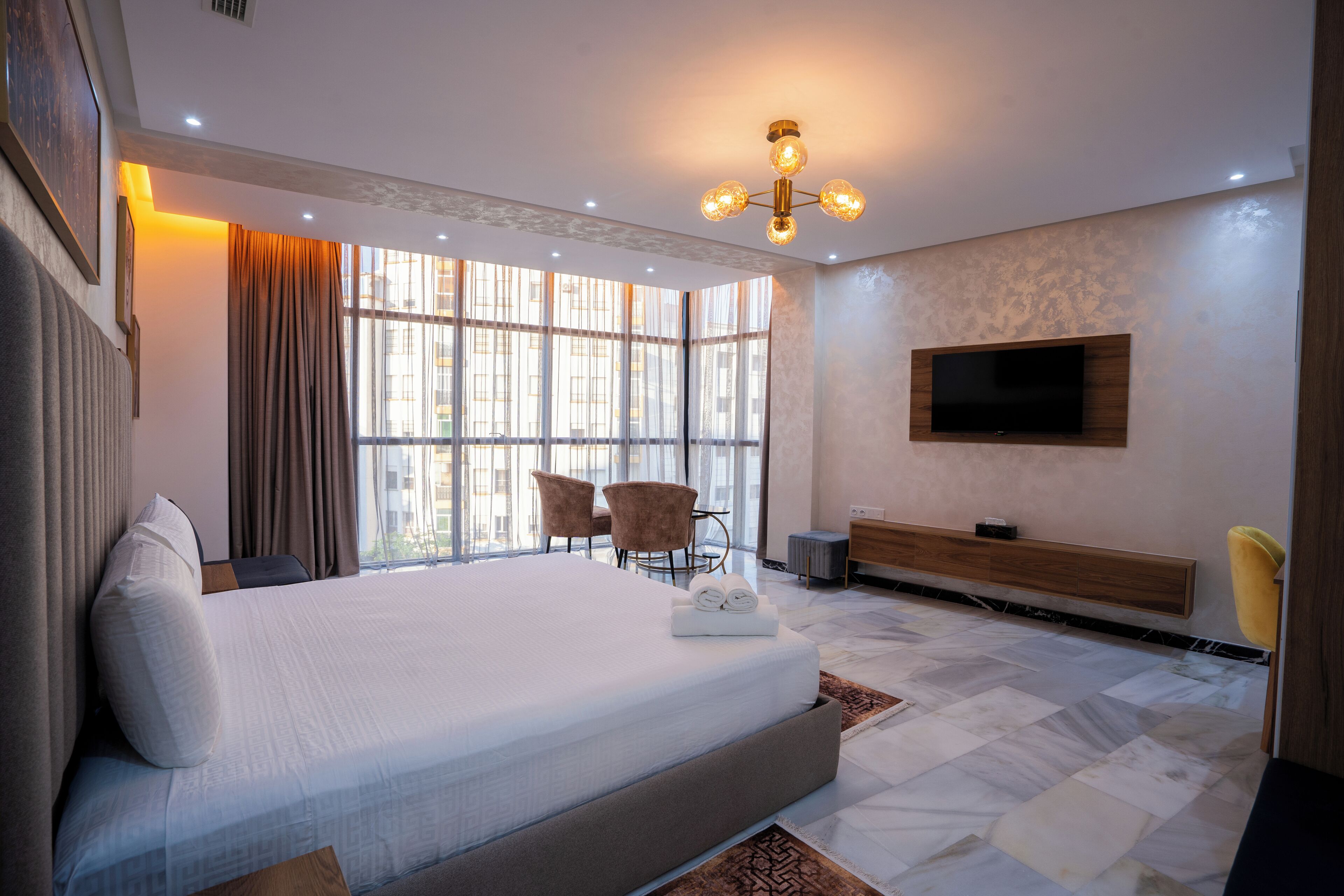Una habitación de hotel moderna con una cama grande, un televisor montado en la pared y un espacio de descanso con sillones.