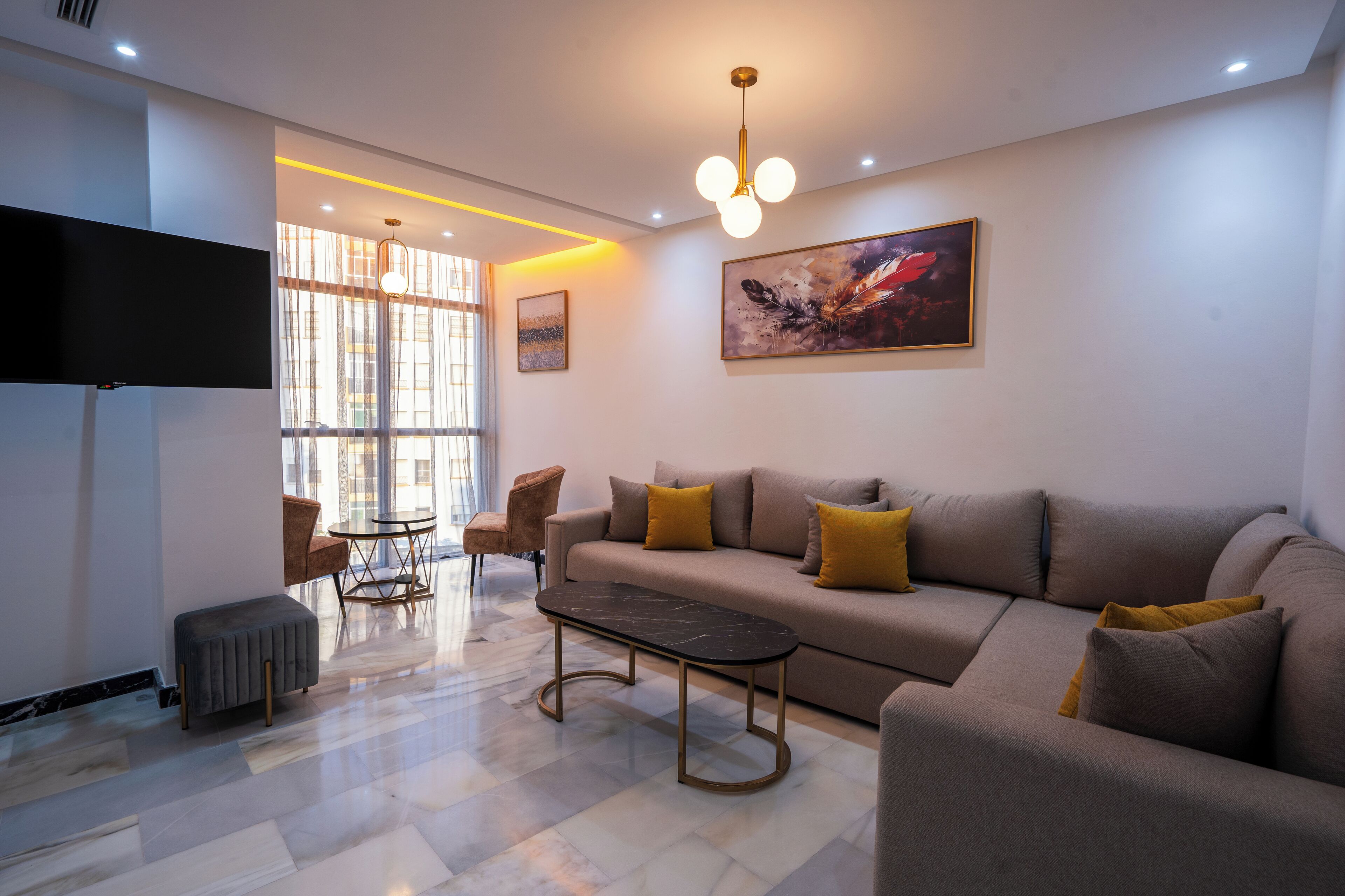 Una sala de estar moderna con un sofá, una mesa de centro y sillones. Hay un televisor de pantalla plana montado en la pared y un cuadro sobre el sofá.