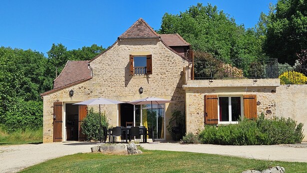 Una casa de piedra con balcón, un comedor con mobiliario exterior y un camino de entrada de grava.