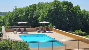 A rectangular pool with a tiled deck, surrounded by lounge chairs and umbrellas, set against a backdrop of dense green trees.