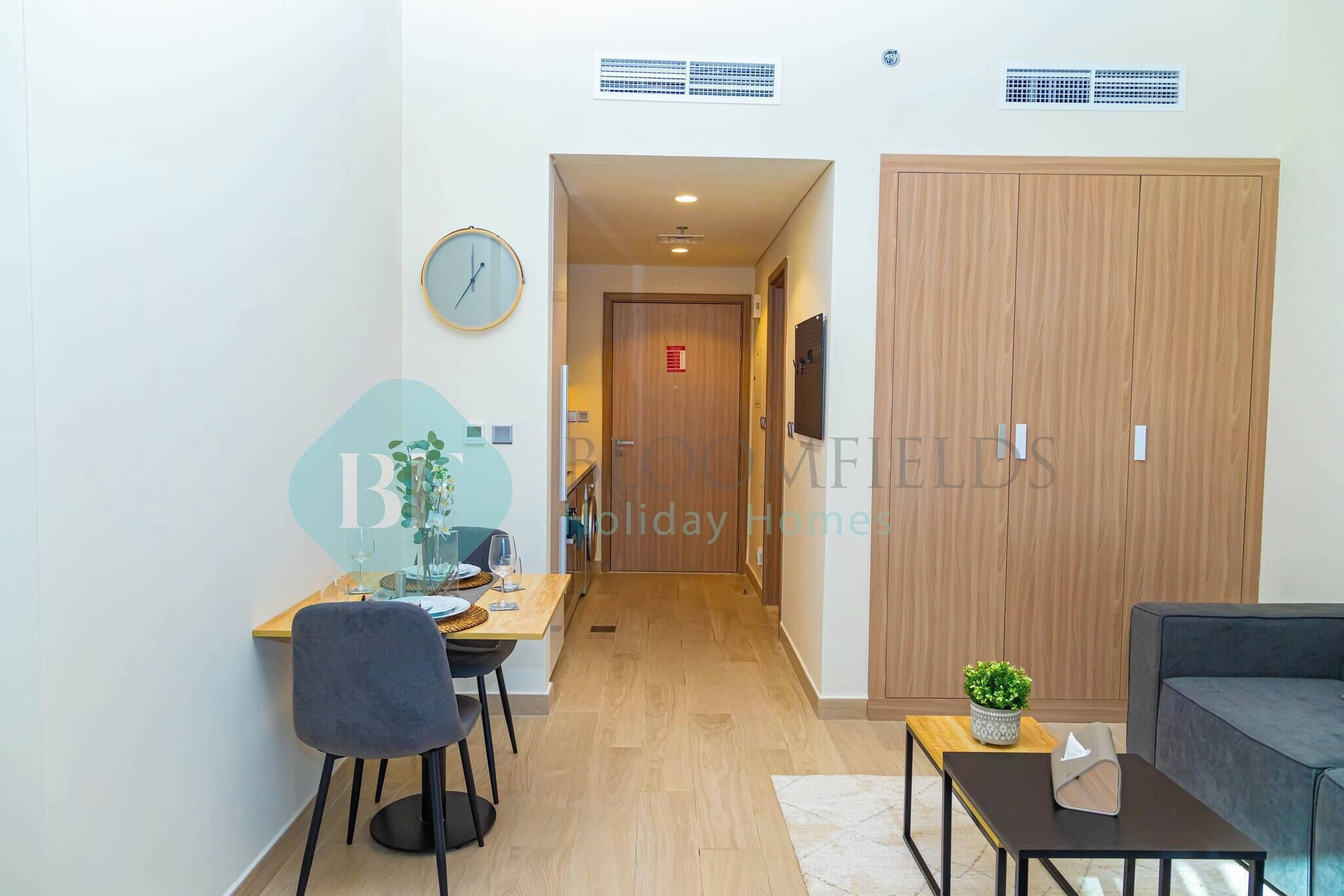 A modern hotel room with a wooden wardrobe, a dining area with a table and chairs, a sofa, and a small potted plant.