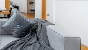 En moderne stue med en grå sofa, et uldtæppe og en sort pude. En trædør fører til et køkken med hvide skabe.
