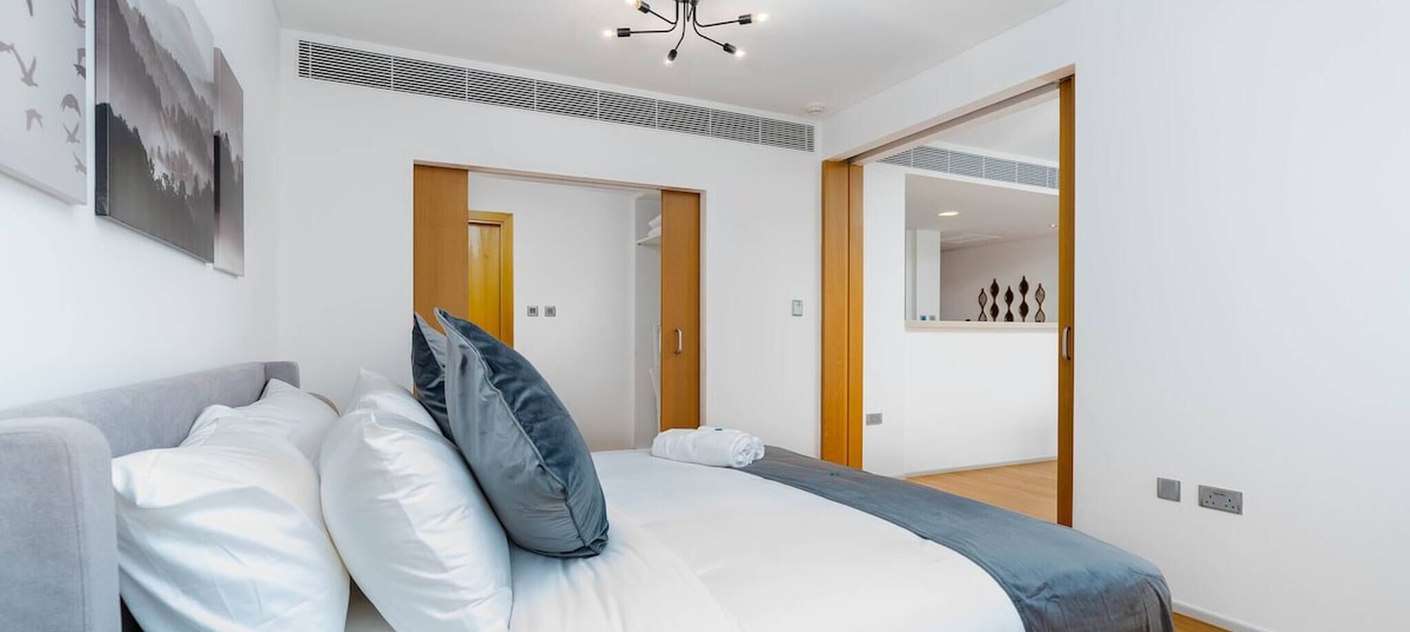 A modern hotel room with a large bed, a grey sofa, and a wooden door leading to a bathroom.