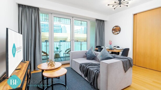 En moderne stue med sofa, et lille bord og et fjernsyn. Der er en balkon med udsigt over bygninger og et spiseområde med bord og stole.