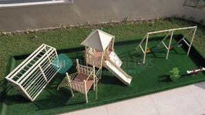 Área de recreação infantil com escorrega, estrutura de escalada e balanços em grama sintética.