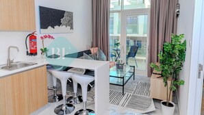 Une cuisine moderne avec un plan de travail blanc, un  évier en acier inoxydable et un extincteur. On y trouve un coin repas avec une table en verre et des tabourets de bar blancs. Une plante en pot est visible dans un coin.