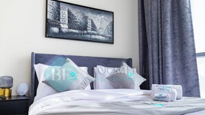 Cama bem arrumada com lençóis e almofadas brancos, um quadro na parede e uma mesa de cabeceira com abajur e um pequeno aparelho.