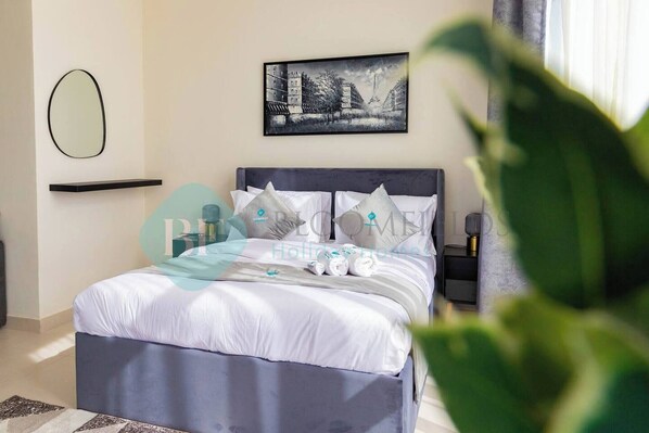 A neatly made bed with white linens and decorative pillows, a black headboard, a bedside table with a lamp, and a framed cityscape picture on the wall.