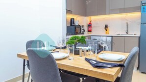 Uma sala de jantar moderna com uma mesa de madeira posta para quatro pessoas, uma pequena planta em vaso e uma cozinha ao fundo com uma máquina de café e um extintor de incêndio.