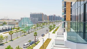 A modern building with a glass facade, a wide street with palm trees, and other high-rise buildings in the background. - Bloomfields STU City View Azizi Riviera Meydan (Dubai)