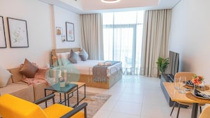 A modern hotel room with a bed, sofa, dining table, and chairs. There are framed pictures on the wall and a window with curtains. - Bloomfields Spacious STU in Thalassa Beach Front (Abu Dabi)