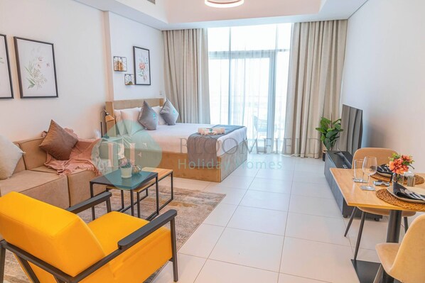 A modern hotel room with a bed, sofa, dining table, and chairs. There are framed pictures on the wall and a window with curtains.