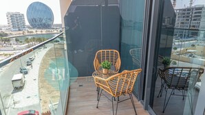 A balcony with wicker chairs and a small table, overlooking a cityscape with modern buildings and a road. - Bloomfields Spacious STU in Thalassa Beach Front (Abu Dabi)
