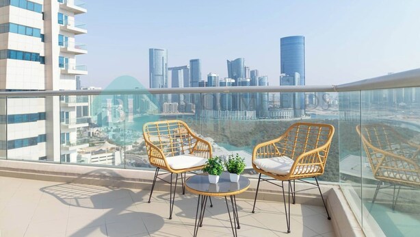 A balcony with two wicker chairs and a small table, overlooking a cityscape with modern buildings. - Bloomfields 1BR Ocean View in OceanScape (Abu Dabi)