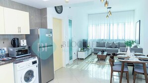 A modern kitchen with a washing machine, a refrigerator, and a microwave. There is a dining area with a table set for two, a sofa, and a coffee table. - Bloomfields 1BR Ocean View in OceanScape (Abu Dabi)
