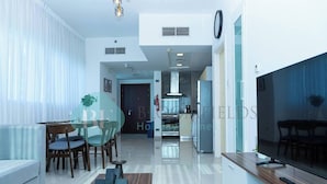 A modern living room with a sofa, dining table, and chairs, a kitchen area with appliances, and a large TV. - Bloomfields 1BR Ocean View in OceanScape (Abu Dabi)