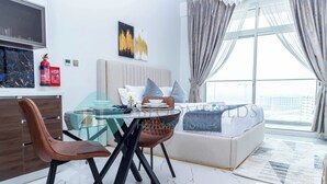 A modern living room with a glass dining table, brown leather chairs, a sofa with a 'Holiday Homes' sign, a large window with curtains, and a fire extinguisher on the wall. - Bloomfields Hidden Gem Studio In Arjan (Dubai)