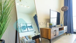 A modern living room with a large mirror, a wooden cabinet, a floor lamp, and a potted plant. - Bloomfields Stylish 1BR Binghatti Heights JVC (Dubai)