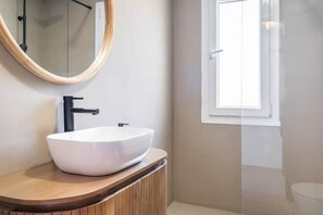 Une salle de bain moderne avec un lavabo sur colonne blanc, un meuble-lavabo en bois, un miroir rond et une fenêtre.