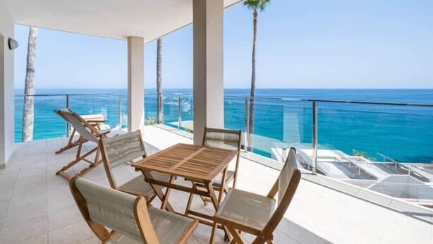 A balcony with a wooden table and chairs, overlooking a clear blue ocean.
