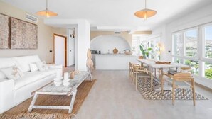 A modern living room with a white sofa, a coffee table, and a dining area with a table and chairs. There is a kitchen in the background with a white countertop and a hanging pot.