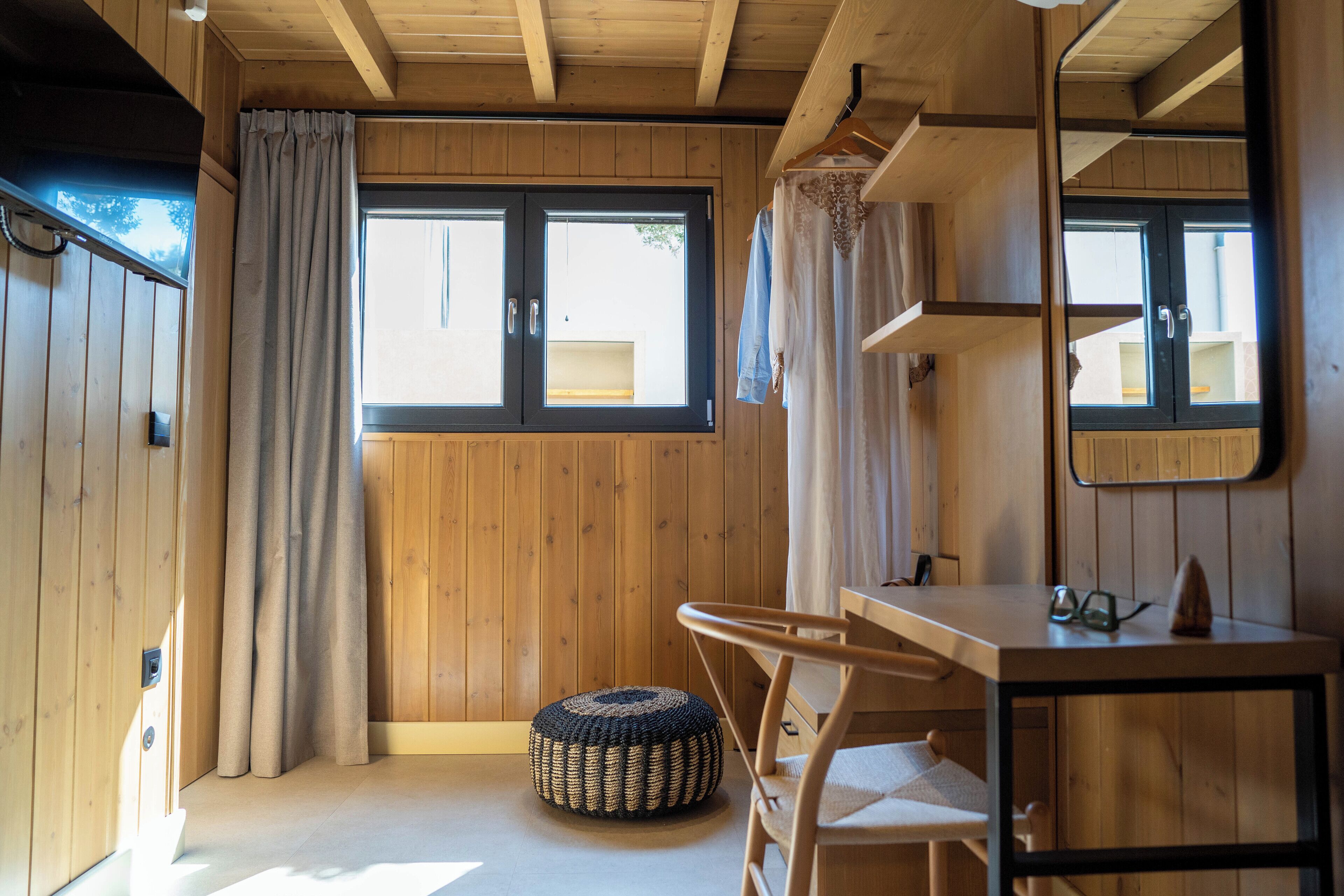 Une pièce en bois avec une grande fenêtre, une chaise, un petit pouf et un miroir au mur.
