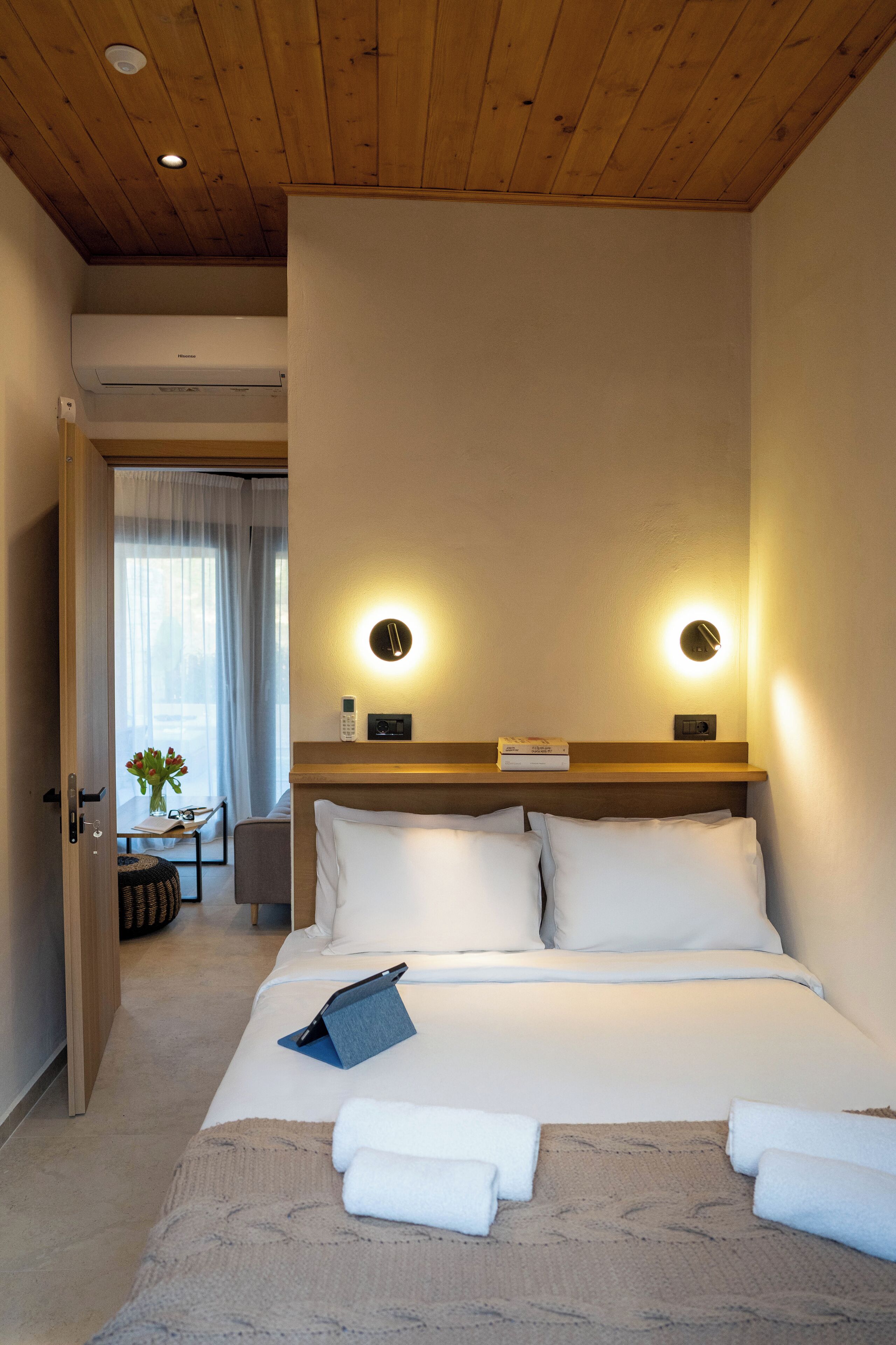A hotel room with a wooden ceiling, a bed with white bedding, two bedside lamps, a bedside table with a phone and a book, and a view of a room with a window and curtains.