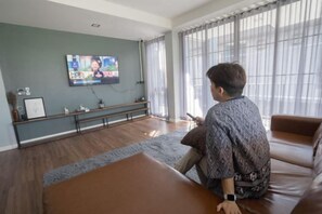 Un hombre sentado en un sofá de cuero marrón frente a un televisor en una habitación con piso de madera, un banco y una ventana con cortinas.
