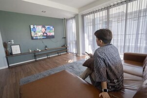 A man sitting on a brown leather couch facing a television in a room with a wooden floor, a bench, and a window with curtains.
