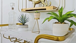 Una encimera de mármol con una bandeja de marco dorado, un decantador de vidrio, una pequeña planta en maceta, una planta más grande en maceta y una escultura de gato dorada sobre una estantería.