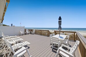Eine Holzterrasse mit weißen Stühlen und Tischen, mit Blick auf einen Strand.