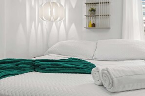 Una cama bien tendida con un edredón blanco, una toalla blanca enrollada y una manta verde.