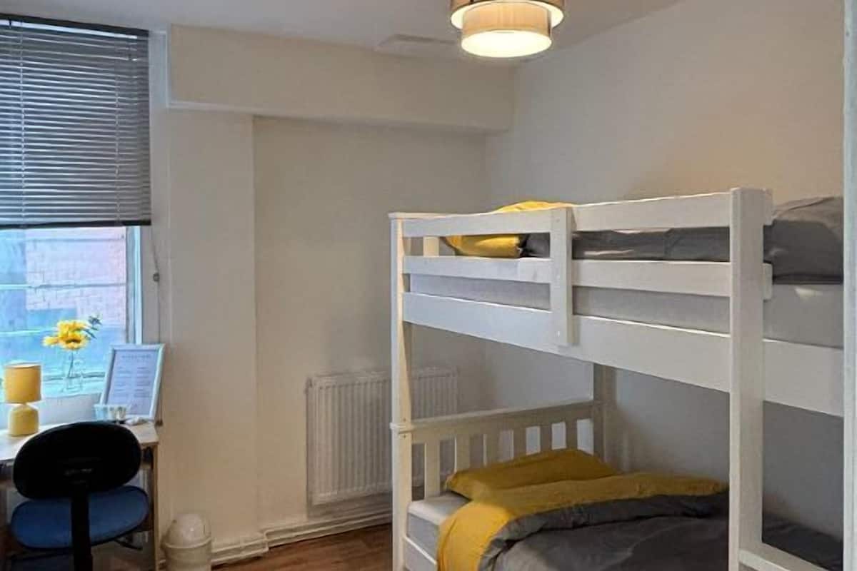 A room with bunk beds, a desk with a chair, a radiator, and a window with blinds.