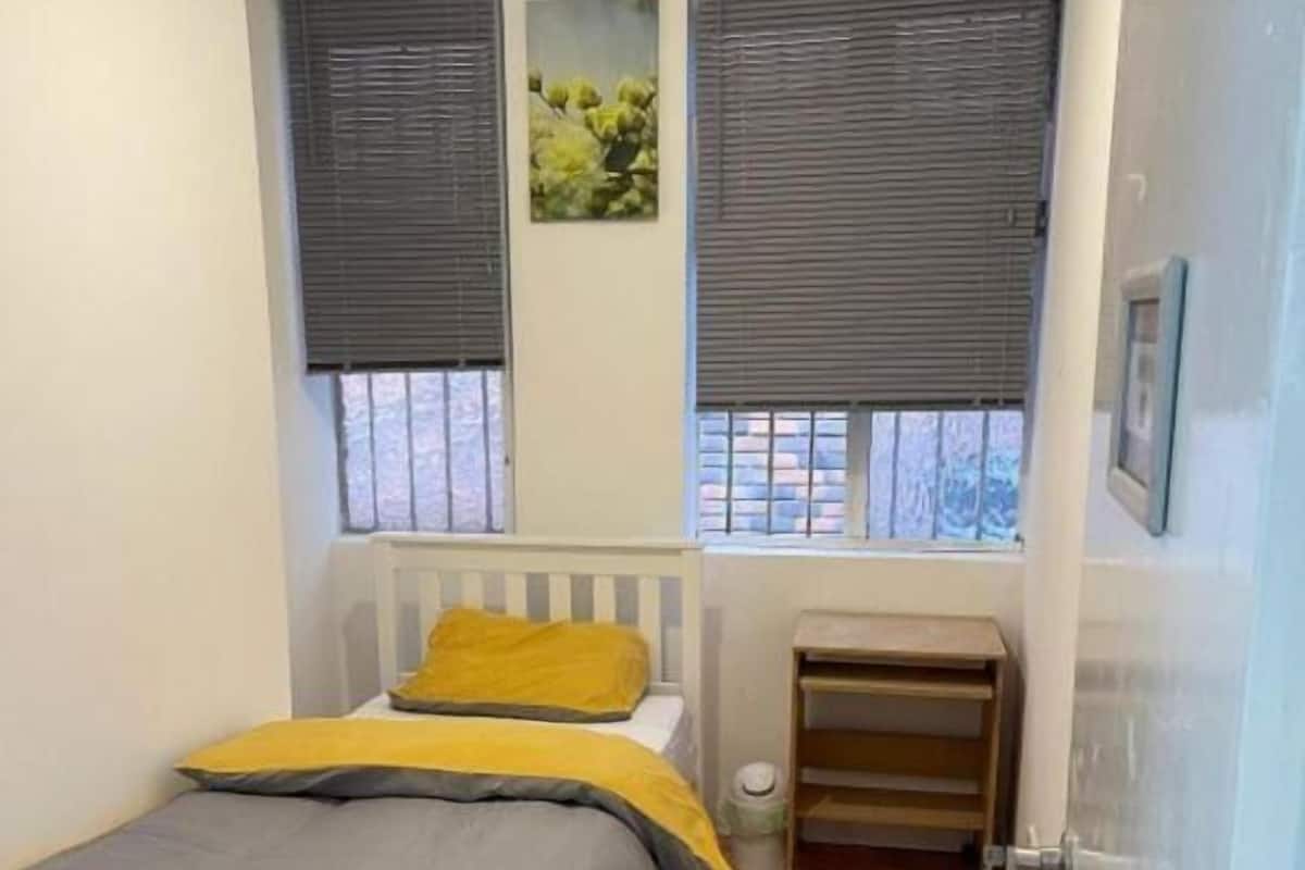 A bedroom with a bed, a bedside table, a window with blinds, and a picture on the wall.
